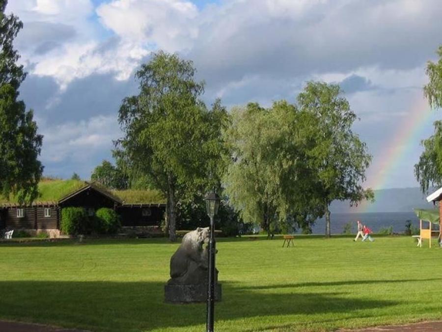 Timmerhus med stor gräsmatta framför och sjö och regnbåge i bakgrunden. 