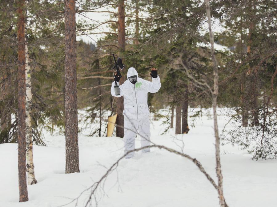 White-clad person with paintball rifle stretched in the air with fir trees around in snowy landscape.