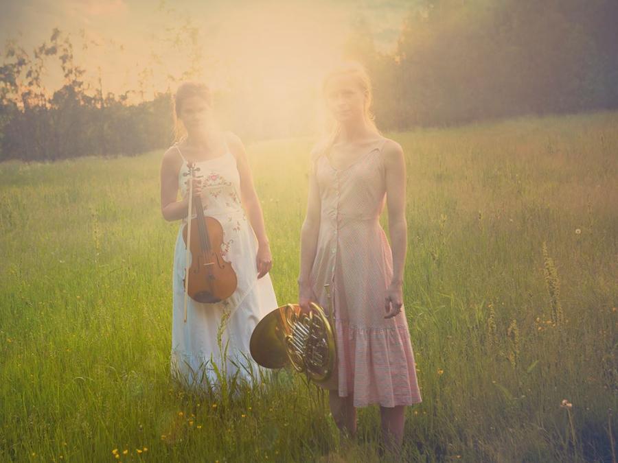 Två kvinnor i klänning på en sommaräng i solnedgång, båda har ett instrument i händerna.