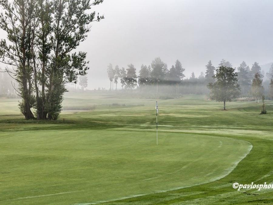 En flagga vid ett golfhål, träd och dimslöjor i bakgrunden.