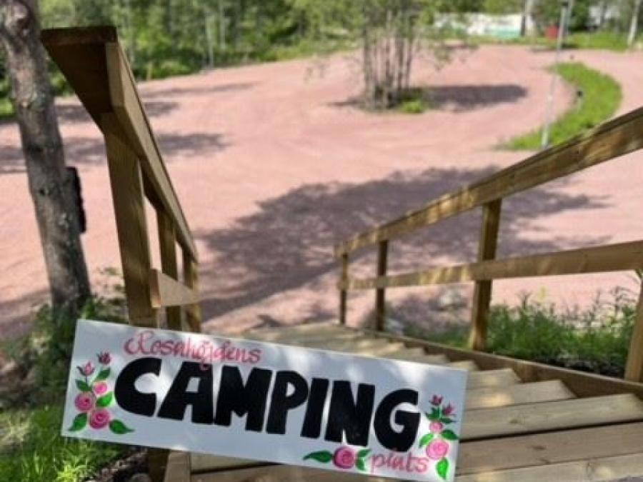 En skylt med texten Rosahöjdens camping plats placerad i en trä trappa, i bakgrunden en grusplan ogiven av träd.