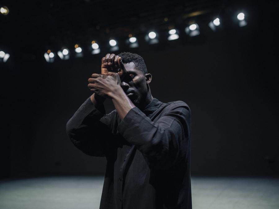 En man i mörk skjorta som står på en scen, håller händerna för ansiktet, spotlights i bakgrunden.