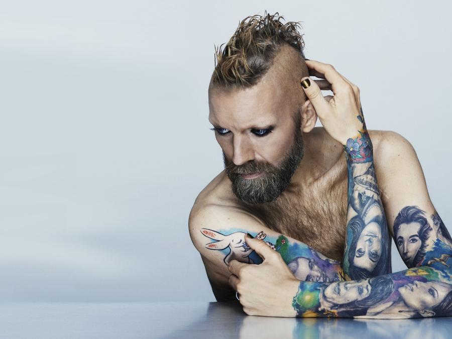En man i skägg och snaggat hår med tatueringar på båda armarna, armarna stöder han på ett bord, blicken snett nedåt.