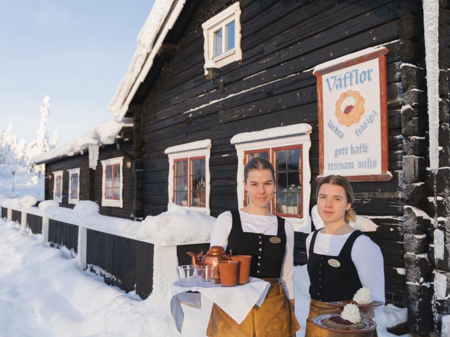 Two ladies stand outside a brown log house with surrounding snow, each holding a tray with coffee cups, a coffee pot and waffles.