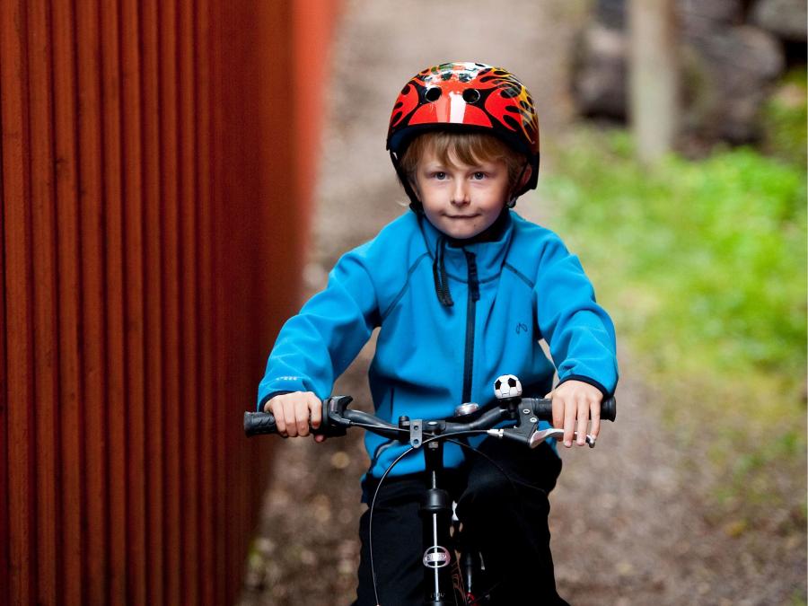 A boy in blue jacket and red helmet biking on a gravel road, red wooden house on the side.