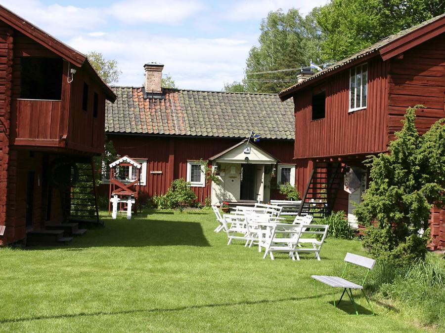 Gårdstun med röda timmerbyggnader i äldre stil, vita trädgårdsmöbler utplacerade på gräset.