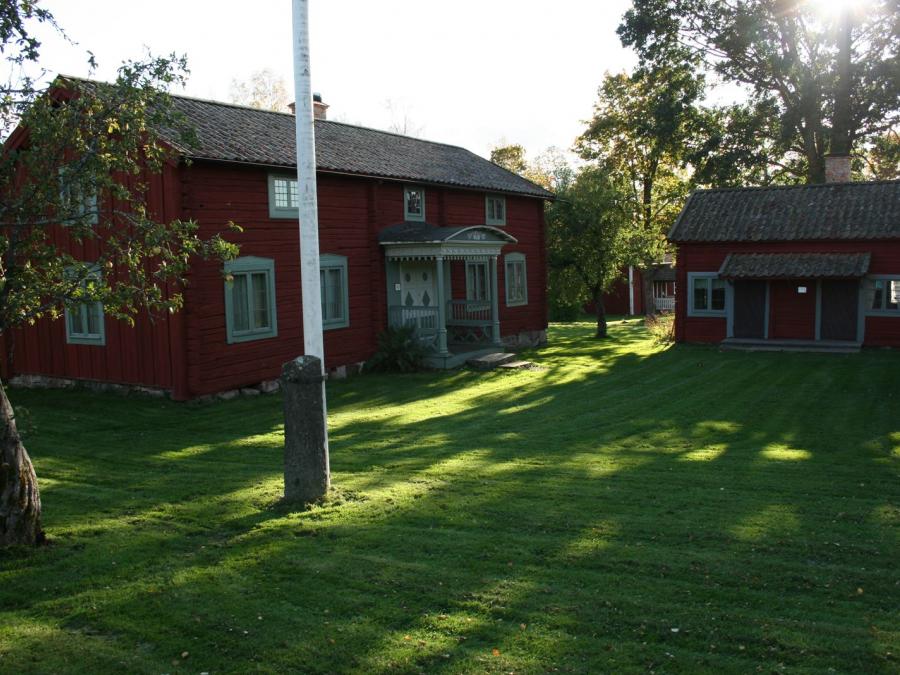 Gräsmatta, nedre delen av en flaggstång, två röda timmerbyggnader, en större i två våningar med farstukvist och en mindre.
