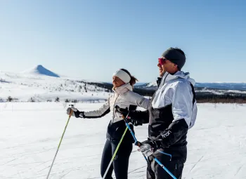 En man och en kvinna i skidkläder, blå himmel, sol och en spetsig bergstopp i bakgrunden.