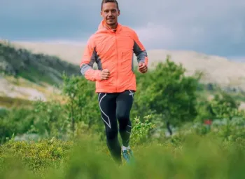 Man springandes i fjällmiljö med orange vindjacka och svarta byxor.