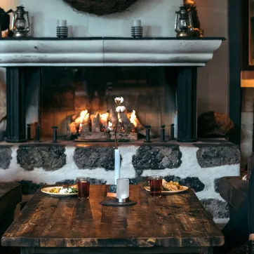 En kvinna i keps sitter vid ett dukat bord framför en brasa.