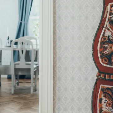 Painted red wooden folk-art piece stands by patterned wallpaper, with a dining table and white chairs in the next room.