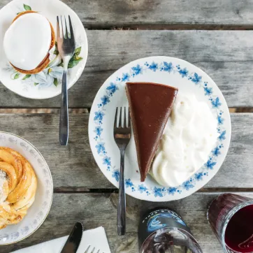 Overhead view of chocolate cake with whipped cream, a cinnamon bun, a meringue, and a red drink on a wooden table.