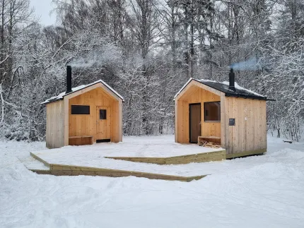 Två små träbyggnader med skorstenar, trall framför, vinterlandskap runtom.