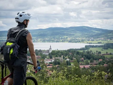 En cyklist som står och tittar ut över en by, sjö och berg i bakgrunden.
