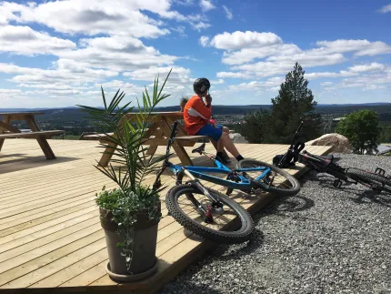 Två cyklar som ligger mot en trätrall, en kille i cykelhjälm som sitter på en bänk, träd, utsikt.