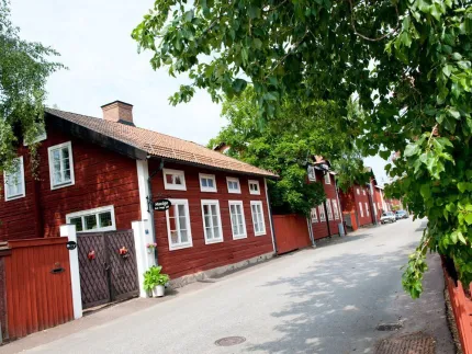 Röd träbyggnad med vita fönster längs en stilla gata, grönskande träd hänger in över vägen i dagsljus.