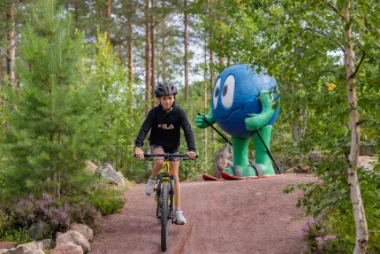 A child biking on a trail, Orsa Grönklitt's mascot in the form of a blueberry in the background.