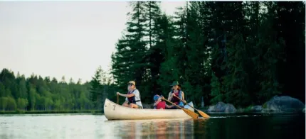 Tre personer paddlar en kanot på en lugn sjö med tät barrskog och några klippor i bakgrunden.