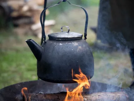En sotig svart kaffepanna hänger över en lägereld med synliga lågor och rök utomhus på gräs.