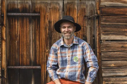 En man i rutig skjorta och svart slokhatt, sitter ner och har en timmervägg i bakgrunden.