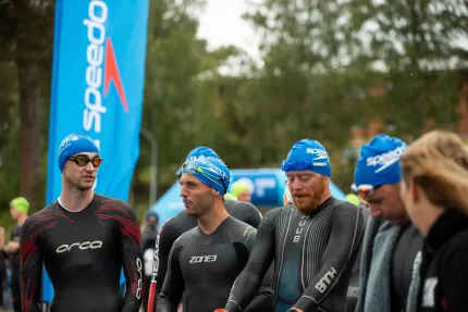 Deltagare i våtdräkt och badmössor, beachflagga och träd i bakgrunden.