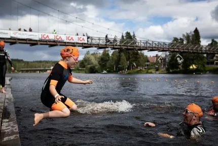 En flicka i våtdräkt och orange badmössa hoppar ner i öppet vatten, intruktör i vattnet, bro i bakgrunden.