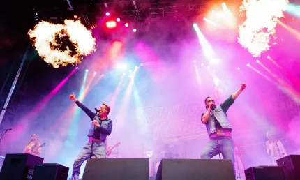 Två män i jeans och jeansväst med varsin mikrofon på en scen, spotlights och eld.