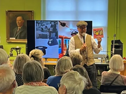 En man i keps med mikrofon som håller upp en bok, publik, bildskärm och symaskin i bakgrunden.