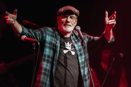 En man i keps och rutig skjorta, en mikrofon framför sig, sträcker ut händerna.