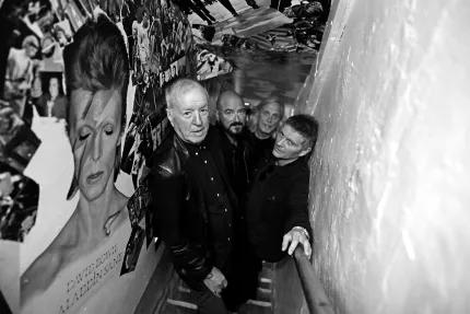 Fyra män som står i en trappa, en vit stenvägg på ena sidan och en målning av David Bowie på den andra.