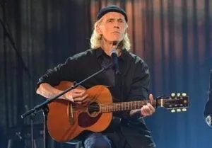 En man sitter på en stol, håller i en gitarr, en miklrofon framför sig, iklädd mörk jacka och skjorta, blå jeans och en svart mössa på huvudet.