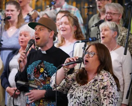 En kör som sjunger, i framkant en man i keps prästkrage och svart tröja med färgglatt mönster och en kvinna i långt mörkt hår och en mönstrad ljus tröja.
