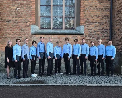 Unga män i blåa skjortor och svarta byxor och en kvinna i svart klänning står på rad framför en tegelbyggnad.