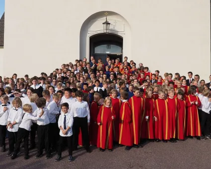 En stor grupp killar framför en kyrka, några i svarta byxor och vita skjortor, några i röda caper med en gul rand.