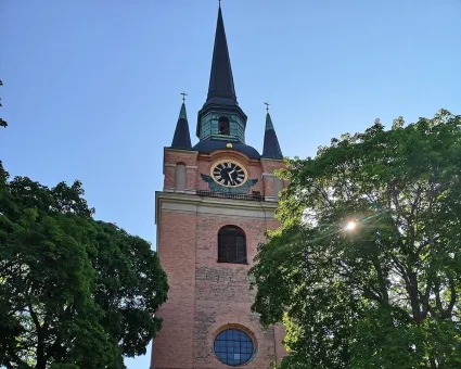 Ett kyrktorn i rött tegel, grönskande träd på sidorna, solen lyser in mellan träden.