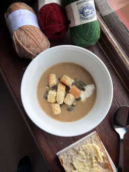 En skål med soppa toppad med brödkrutonger och pumpakärnor, brevid en bit bröd en sked och garnnystan.