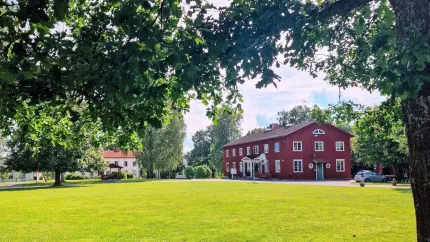 Ett rött tvåvåningshus i trä, en vit byggnad skymtar bakom trädet, trädgrenar och en stor gräsmatta i framkant.