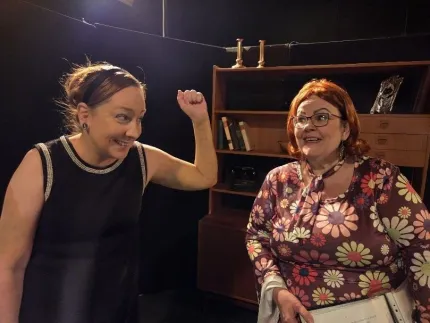 Två kvinnor, en i uppsatt hår ikäff ärmlös svart klänning håller upp ena handen, den andra kvinnan har glasögon och är  iklädd blommig klänning.