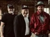 Tre personer står nära varandra inomhus; två i mörka tröjor och en i röd jacka samt keps, träbakgrund.