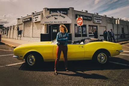 En kvinna med armarna i kors står mitt på en gata framför en gul sportbil.