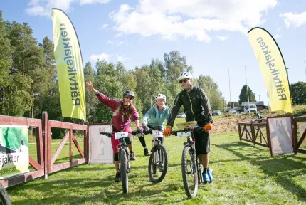 Tre cyklister med hjälm leder mountainbikes över gräs vid en startgrind med flaggor, träd och blå himmel..