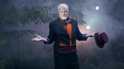 En man i mörk kavaj och orange tröja står i dimmig barrskog med utsträckta armar och håller en rutig hatt..