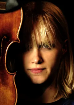 Närbild av en violin i trä bredvid en person med ljust hår, mot mörk bakgrund i varmt ljus..