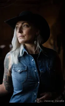 En kvinna med långt ljust hår  iklädd jeansväst och en svart cowboyhatt på huvudet.