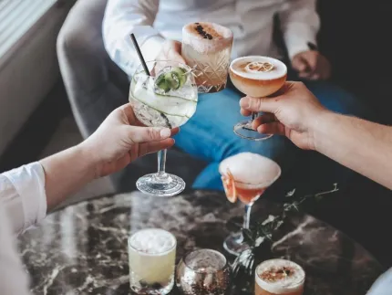 Händer som skålar med cocktails i olika glas ovanför ett marmorbord, med fler drinkar och citrusdekorationer..