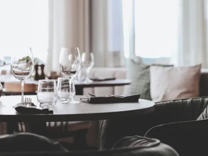 Runt bord med vinglas, vattenglas och bestick i en ljus restaurang med soffkuddar och stora fönster..