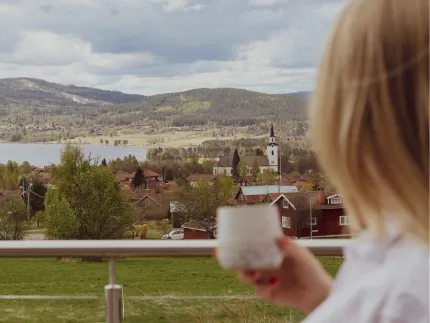 En person med en kopp i handen ser ut över en by med kyrktorn, sjö och skogklädda berg under molnig himmel..