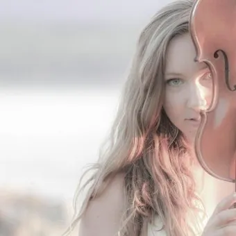 Närbild av en person med långt ljust hår mot en ljus, suddig bakgrund som liknar strand eller vatten..