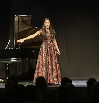 En person i lång mönstrad klänning står på en scen bredvid en flygel, med publik i förgrunden..