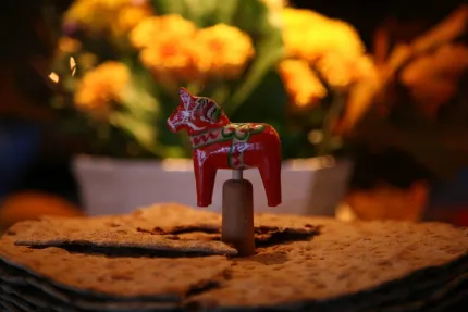 Röd dalahäst på pinne står i en stapel knäckebröd, med suddiga gula blommor i bakgrunden..